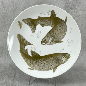 May include: A white ceramic plate with a gold design of two trout swimming in opposite directions. The plate has a simple, elegant design.