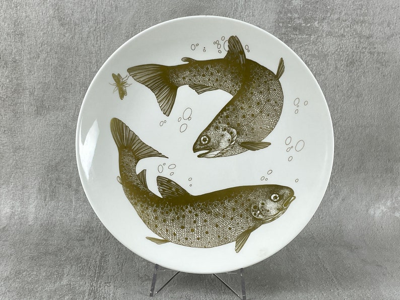 May include: A white ceramic plate with a gold design of two trout swimming in opposite directions. The plate has a simple, elegant design.