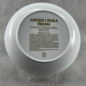 May include: A white ceramic plate with a gold border and text in Swedish. The text reads "ARTER I FARA &Ouml;RING Nr 11 i en serie samlartallrikar i benporslin, som tillverkats i en mycket begr&auml;nsad upplaga. Dekoren &auml;r i guld Design: Paul Hoff &Ouml;RING (Salmo trutta) Liksom sin n&auml;ringsringen bredd over hela Sverige. Bagge arterna kr&auml;ver str&ouml;mmande vatten orleken. Av bring brekommer former som vandrar ut havet eller til de stora inajama f&ouml;r att som k&ouml;nsmogna Aterv&auml;nda i sina uppv&auml;tomr&aring;denez haradring, Oring). Andra former er station&auml;ra, dva lever hela livet i samma begr&auml;nsade vattensystem (h&auml;ck&ouml;ring Oringen &auml;r mycket variationsrik och m&aring;nga olika bestand har utbildats. Genom bi a dammbyggnader och stregle ringar, f&ouml;rsuming och kalkning samt fororeningar har ra denna situation manga h&aring;ll. En betydande arbete l&auml;ggs ned p&aring; att f&ouml;rba B&auml;ck&ouml;ringama (Salmo trutta fario p&aring; talkan f&aring;r repre sentera olika atera bring"