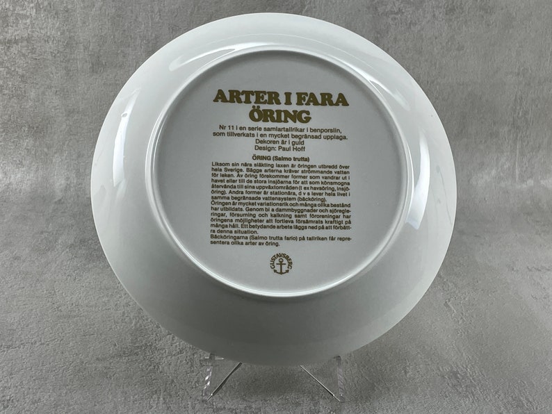 May include: A white ceramic plate with a gold border and text in Swedish. The text reads "ARTER I FARA &Ouml;RING Nr 11 i en serie samlartallrikar i benporslin, som tillverkats i en mycket begr&auml;nsad upplaga. Dekoren &auml;r i guld Design: Paul Hoff &Ouml;RING (Salmo trutta) Liksom sin n&auml;ringsringen bredd over hela Sverige. Bagge arterna kr&auml;ver str&ouml;mmande vatten orleken. Av bring brekommer former som vandrar ut havet eller til de stora inajama f&ouml;r att som k&ouml;nsmogna Aterv&auml;nda i sina uppv&auml;tomr&aring;denez haradring, Oring). Andra former er station&auml;ra, dva lever hela livet i samma begr&auml;nsade vattensystem (h&auml;ck&ouml;ring Oringen &auml;r mycket variationsrik och m&aring;nga olika bestand har utbildats. Genom bi a dammbyggnader och stregle ringar, f&ouml;rsuming och kalkning samt fororeningar har ra denna situation manga h&aring;ll. En betydande arbete l&auml;ggs ned p&aring; att f&ouml;rba B&auml;ck&ouml;ringama (Salmo trutta fario p&aring; talkan f&aring;r repre sentera olika atera bring"