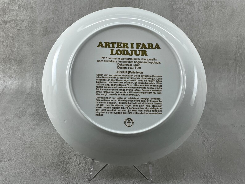 May include: A white ceramic plate with a gold border and text in Swedish. The text reads "Arter i Fara Lodjur" (Species in Danger Lynx) and includes information about the European wildcat and the lynx. The plate is part of a series of collector's plates.