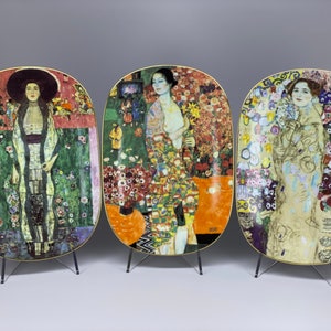 May include: Three oval-shaped ceramic plates with gold trim, each featuring a different painting of a woman in a colorful floral setting. The plates are displayed on individual metal stands.