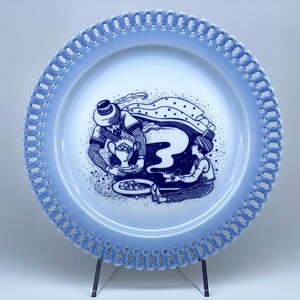 Puede incluir: Un plato de porcelana azul y blanco con un borde de encaje decorativo. El plato presenta una escena de dos figuras en azul y blanco, una sosteniendo una taza y la otra una pistola.
