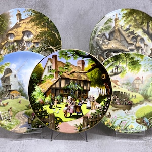 Puede incluir: Conjunto de cinco platos decorativos con ilustraciones coloridas de pintorescas casas de campo inglesas y escenas campestres. Cada plato representa una escena diferente, incluyendo un molino de viento, una casa de campo con techo de paja y un grupo de niños jugando en un jardín.