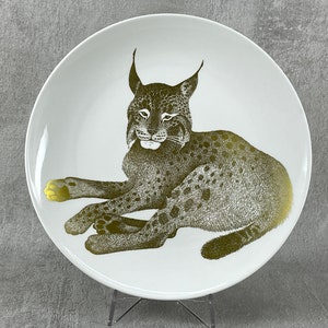 May include: A white ceramic plate with a gold-colored illustration of a lynx lying down. The lynx has a spotted pattern and is facing the viewer.