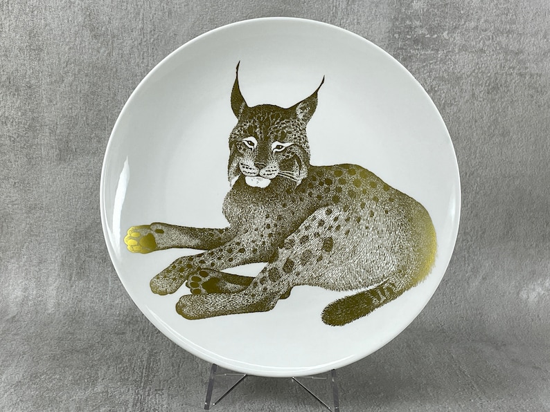 May include: A white ceramic plate with a gold-colored illustration of a lynx lying down. The lynx has a spotted pattern and is facing the viewer.