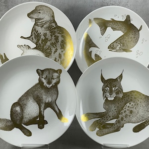 May include: Set of four white ceramic plates with gold accents. Each plate features a different animal: a seal, a fish, a lynx, and a fox.