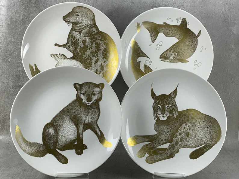 May include: Set of four white ceramic plates with gold accents. Each plate features a different animal: a seal, a fish, a lynx, and a fox.