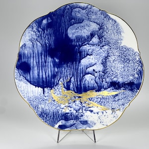 Puede incluir: Un plato de cerámica blanco con un diseño azul y dorado. El plato tiene un borde festoneado y presenta un diseño de pájaro estilizado en dorado.