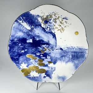 Puede incluir: Un plato de cerámica blanco con un diseño azul y dorado. El plato presenta una escena de paisaje con una montaña, agua y nenúfares. El plato tiene un borde festoneado y está en un soporte.