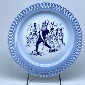 Puede incluir: Un plato de cerámica azul y blanco con un borde decorativo. El plato presenta una escena de un rey en procesión con otras figuras. El plato está en un soporte.