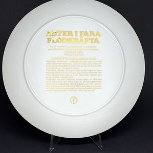 May include: A white ceramic plate with a gold border and text that reads "Arter i fara Flodkrafta" Nr 13 i en serie med motiv av hotade arter, som tillverkats av en svensk designstudio. Design: Paul Folt.