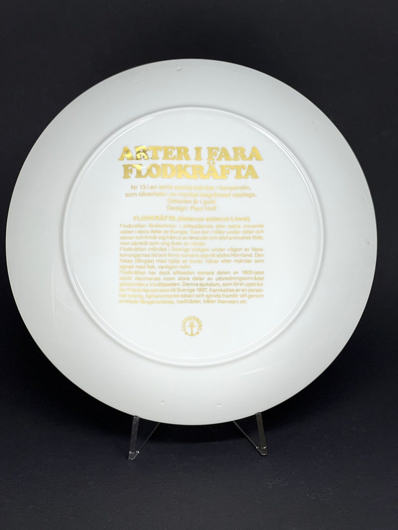 May include: A white ceramic plate with a gold border and text that reads "Arter i fara Flodkrafta" Nr 13 i en serie med motiv av hotade arter, som tillverkats av en svensk designstudio. Design: Paul Folt.