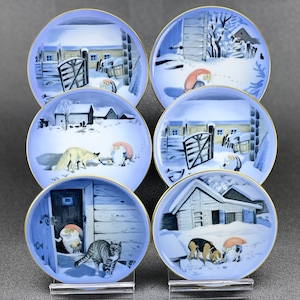 Könnte beinhalten: Satz von sechs blauen und weißen Keramik-Untersetzern mit Winterszenen. Jeder Untersetzer zeigt ein anderes Tier, darunter einen Fuchs, eine Katze, einen Hund und einen Gnom. Die Untersetzer sind mit einem goldenen Rand verziert.