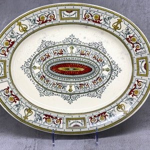 Puede incluir: Fuente ovalada para servir con fondo crema y diseño decorativo ornamentado. El diseño presenta un óvalo central con un patrón rojo y dorado, rodeado de intrincados bordes en oro, rojo y verde.