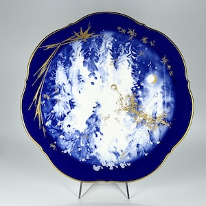 Puede incluir: Un plato de porcelana azul y blanco con un borde dorado y un diseño de árboles y una luna en el centro. El plato está sobre un soporte blanco.