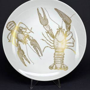 May include: A white ceramic plate with a gold design of two crayfish. The crayfish are detailed with a stippled pattern.