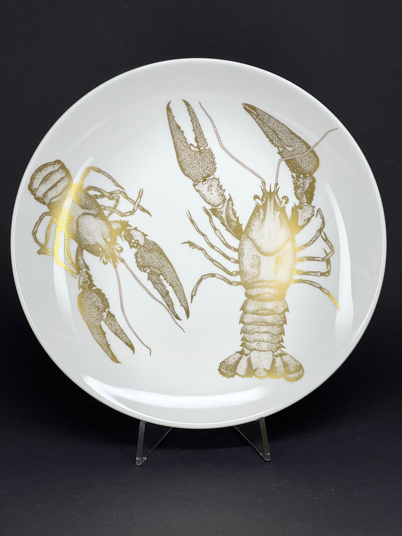 May include: A white ceramic plate with a gold design of two crayfish. The crayfish are detailed with a stippled pattern.