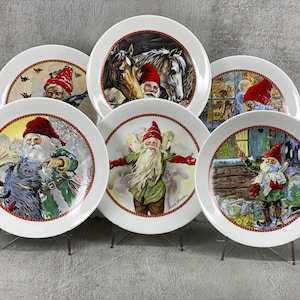 May include: Set of six white ceramic plates with colorful illustrations of gnomes and winter scenes. Each plate features a different gnome with a red hat and a white beard. The plates are perfect for adding a festive touch to your holiday table.