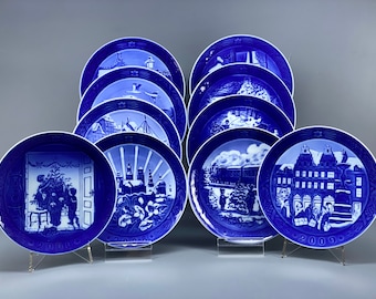 May include: A collection of ten blue and white Royal Copenhagen Christmas plates. The plates feature various scenes of Copenhagen, Denmark, including a Christmas tree, a train, and a church. The plates are numbered 2000 through 2009.