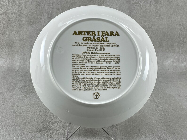 May include: A white ceramic plate with a gold border and text in Swedish. The text describes the gray seal, a species of seal found in the Baltic Sea. The plate is part of a series of collector's plates.