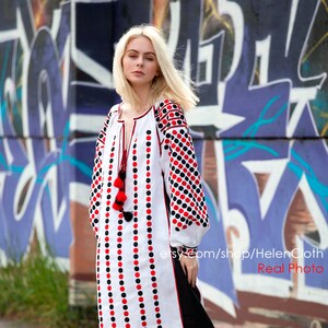 May include: A white linen dress with black and red polka dots. The dress has long sleeves and a round neckline. The dress is embroidered with a floral pattern. The model is wearing the dress with black heeled sandals. The dress is photographed against a graffiti wall.