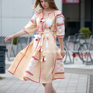 May include: A woman wearing a beige linen dress with white and red embroidery. The dress has a fitted bodice and a flowing skirt. The woman is wearing beige heels and has her hair down.