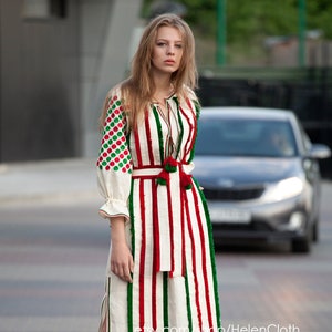 May include: A white linen dress with red and green stripes and embroidered details. The dress has a red sash with tassels and a side slit. The model is wearing the dress with nude heels.