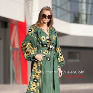 Pode incluir: Um vestido de linho verde com bordados pretos, brancos e dourados. O vestido tem manga comprida, decote redondo e cinto. A modelo está usando óculos de sol e saltos bege.