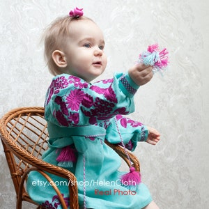May include: A young child wearing a turquoise dress with pink and purple embroidery sits on a wicker chair. The dress has tassels on the sleeves and bottom hem. The child is holding a pink and blue pom-pom.