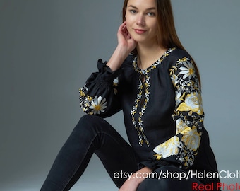 May include: A black linen blouse with white and yellow floral embroidery. The blouse has long sleeves and a round neckline. The model is wearing black pants and is sitting on a white background. etsy.com/shop/HelenCloth Real Photo