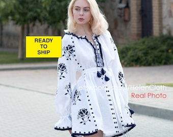 May include: White linen dress with blue embroidered floral pattern. The dress has a round neckline, long sleeves, and a fitted waist. The model is wearing white sneakers.