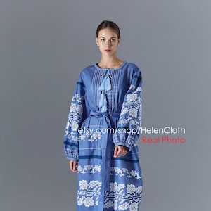 Può includere: Un lungo abito di lino blu con motivi floreali ricamati in bianco. L'abito ha una scollatura rotonda, maniche lunghe e una cintura. L'abito ha un orlo a festoni. La modella indossa sandali marroni.