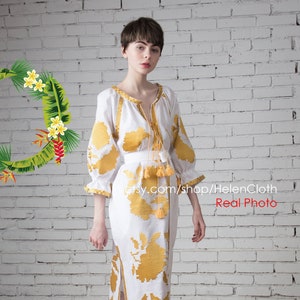 May include: A white linen dress with yellow embroidered floral patterns. The dress has a long skirt and three-quarter sleeves. The model is wearing white sandals.