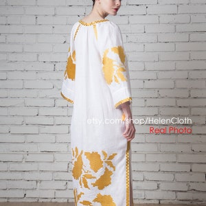 May include: A white linen dress with gold embroidery of floral patterns. The dress has a long, flowing skirt and a loose-fitting bodice. The model is wearing the dress and standing in front of a white brick wall. The text "etsy.com/shop/HelenCloth Real Photo" is visible at the bottom of the image.