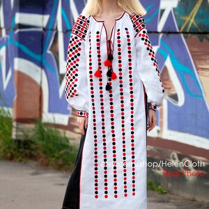 May include: A white linen dress with black and red embroidered polka dots. The dress has a long, flowing skirt and a fitted bodice. The model is wearing the dress with black sandals.