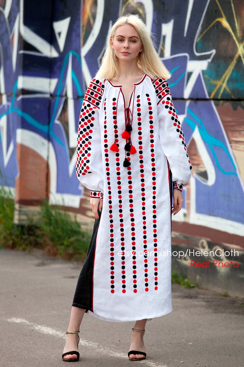 May include: A white linen dress with black and red embroidered polka dots. The dress has a long, flowing skirt and a fitted bodice. The model is wearing the dress with black sandals.