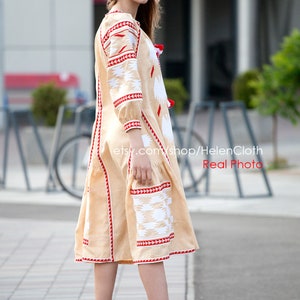 May include: A beige linen dress with red and white embroidery. The dress has a long, flowing skirt and a fitted bodice. The model is wearing the dress with a pair of nude heels.