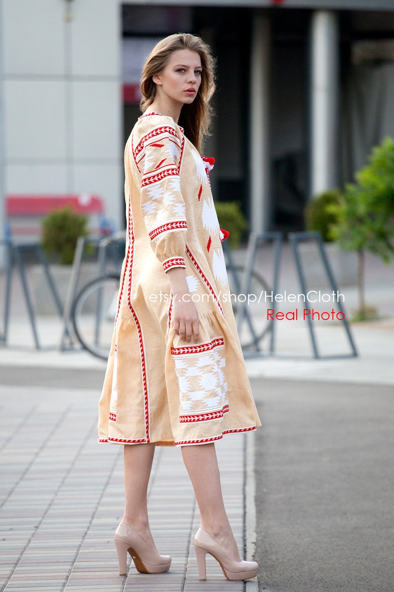 May include: A beige linen dress with red and white embroidery. The dress has a long, flowing skirt and a fitted bodice. The model is wearing the dress with a pair of nude heels.