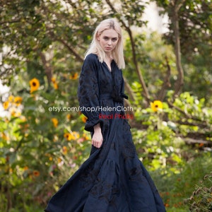 May include: A woman wearing a long, dark blue embroidered dress with a black belt. The dress has a V-neckline and long sleeves. She is standing in a garden with green foliage and yellow flowers. The text "etsy.com/shop/HelenCloth Real Photo" is visible on the image.