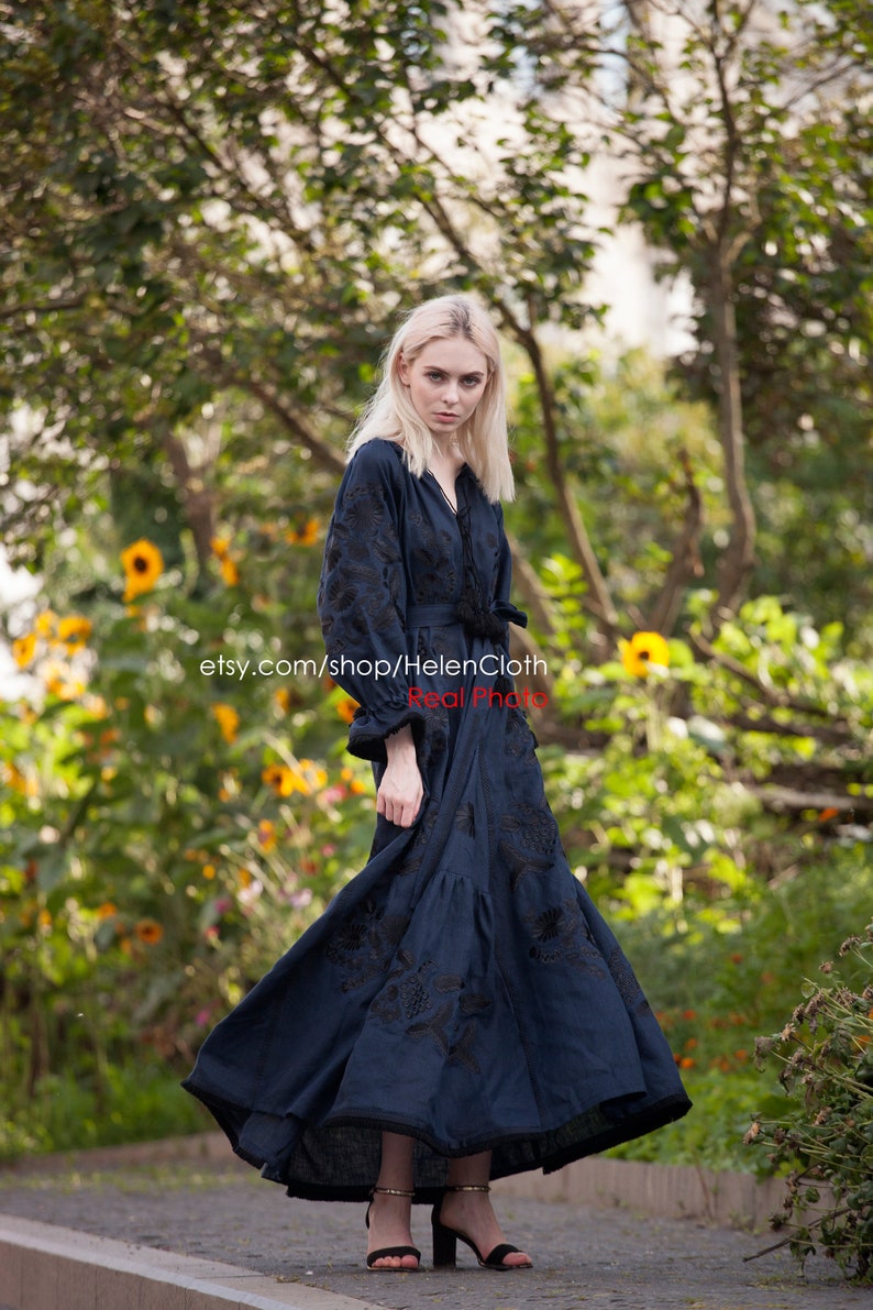 May include: A woman wearing a long, dark blue embroidered dress with a black belt. The dress has a V-neckline and long sleeves. She is standing in a garden with green foliage and yellow flowers. The text "etsy.com/shop/HelenCloth Real Photo" is visible on the image.