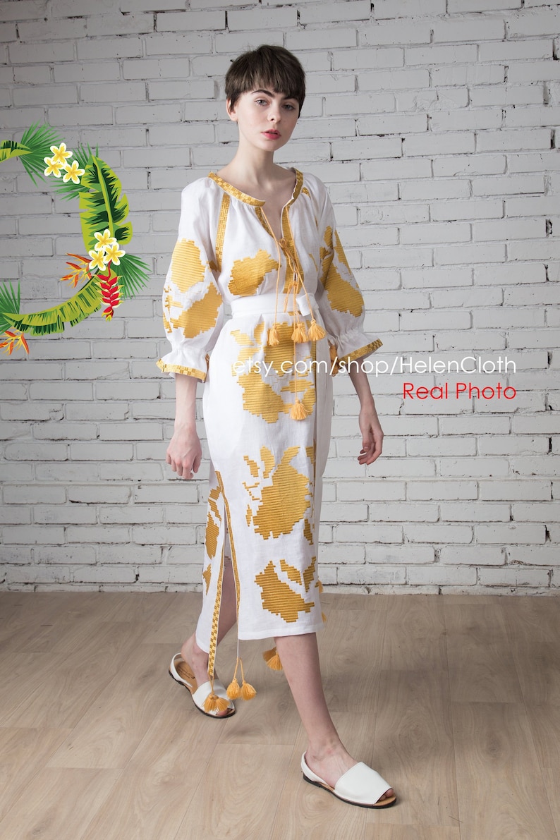 May include: A white linen dress with yellow embroidered floral patterns. The dress has a long skirt and three-quarter sleeves. The model is wearing white sandals.