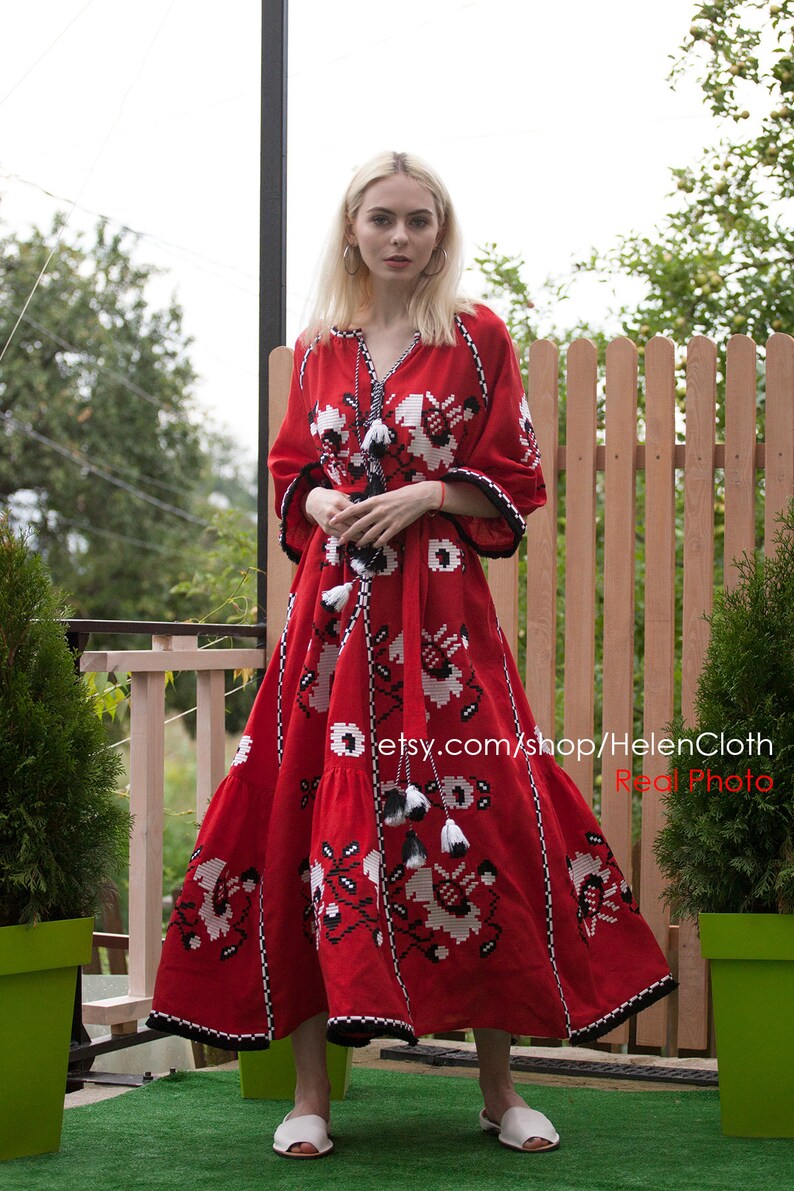 May include: A red maxi dress with white and black embroidery. The dress has a V-neckline and long sleeves. The model is wearing white sandals. The dress is made of linen and has a relaxed fit. The dress is perfect for a summer wedding or a special occasion.  etsy.com/shop/HelenCloth Real Photo