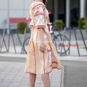 May include: A woman wearing a beige embroidered dress with red and white accents. The dress has a fitted bodice and a flowing skirt. The woman is standing on a sidewalk with a blurred background.