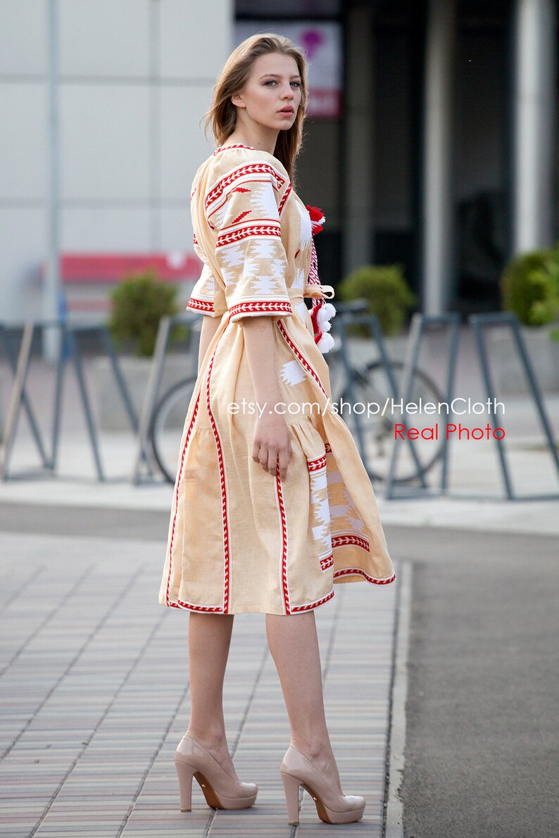 May include: A woman wearing a beige embroidered dress with red and white accents. The dress has a fitted bodice and a flowing skirt. The woman is standing on a sidewalk with a blurred background.