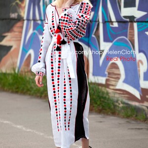 May include: White linen dress with red and black polka dot embroidery. The dress has a long, flowing skirt and a fitted bodice. The model is wearing the dress with black heeled sandals. The dress is photographed against a graffiti wall.