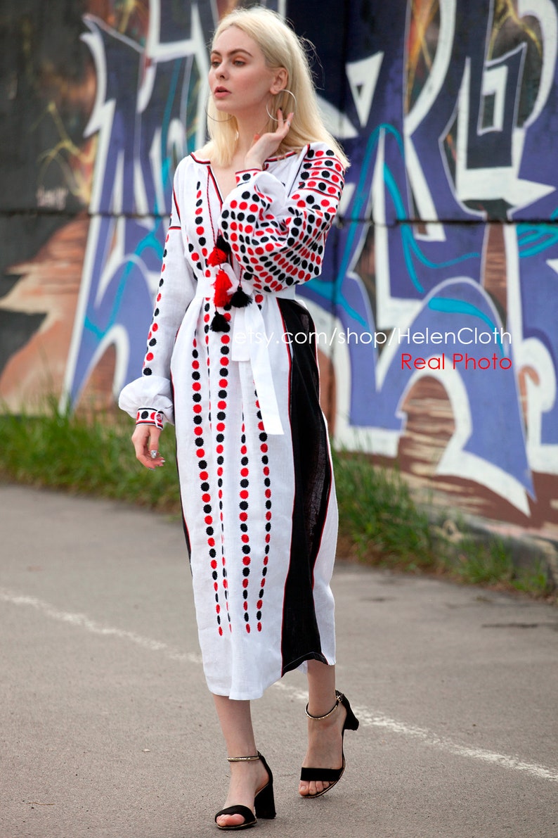 May include: White linen dress with red and black polka dot embroidery. The dress has a long, flowing skirt and a fitted bodice. The model is wearing the dress with black heeled sandals. The dress is photographed against a graffiti wall.