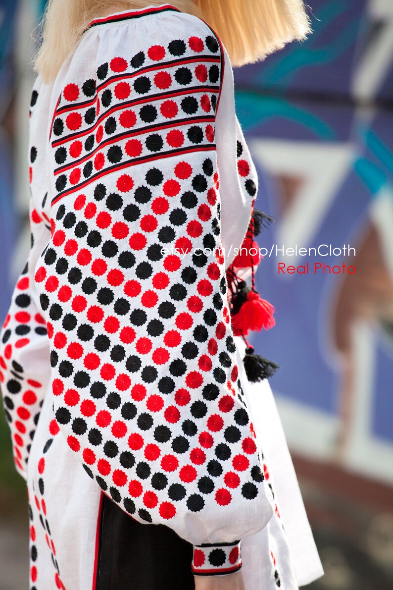 May include: White linen blouse with red and black embroidered polka dots. The embroidery is a traditional Ukrainian design. The blouse has long sleeves and a round neckline.