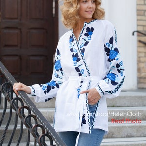 May include: A white linen blouse with blue and black embroidered floral designs. The blouse has a V-neckline and a tie belt. The model is wearing blue jeans and is standing on a set of stairs.