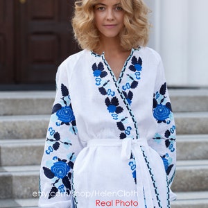 May include: White linen blouse with blue and black floral embroidery. The blouse has a V-neckline and a tie waist. The model is wearing blue jeans. "etsy.com/shop/HelenCloth Real Photo"